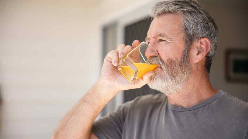An older man with gray hair and a beard drinks orange juice from a glass indoors, wearing a gray t-shirt.