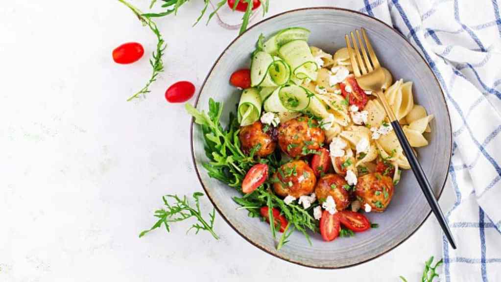 A bowl of pasta salad with meatballs, arugula, cherry tomatoes, cucumber, and feta cheese on a light surface.