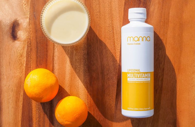 A bottle of Mama liposomal multivitamin next to two oranges and a bowl of juice on a wooden surface.