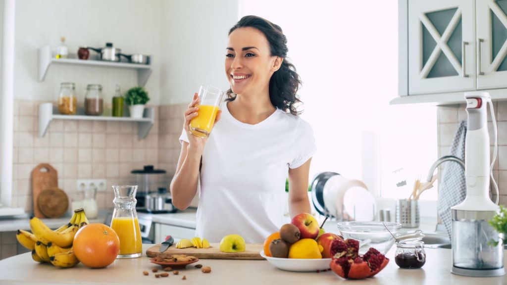 Woman in a white shirt drinking orange juice while standing in a kitchen with various fruits and a juicer on the counter.
