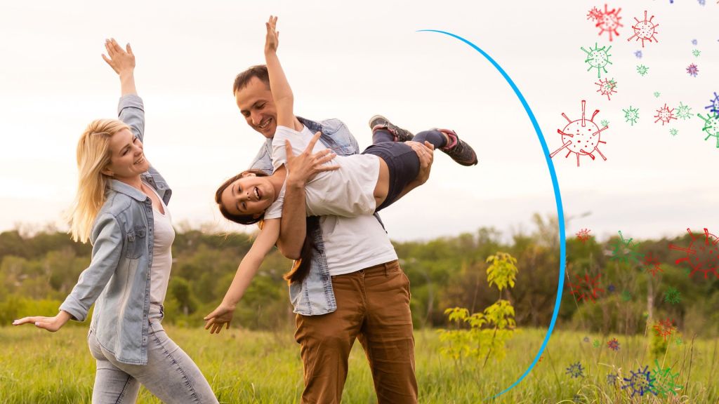 A smiling man holds a laughing child while a woman stands beside them with arms raised in a green field. Colorful virus illustrations are overlaid on the right side of the image.