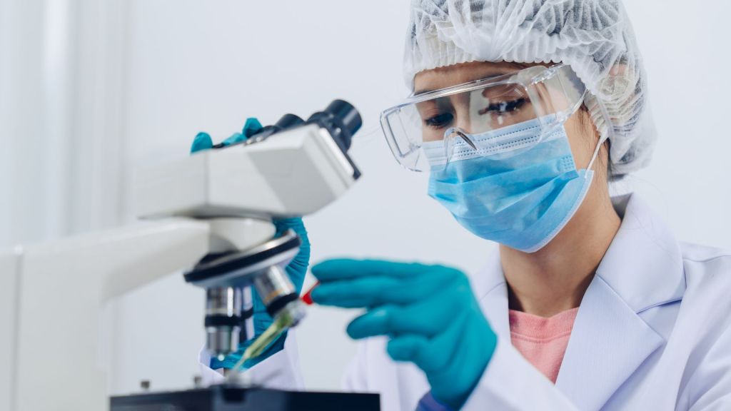 A researcher wearing a lab coat, gloves, hairnet, mask, and safety goggles examines a sample under a microscope.