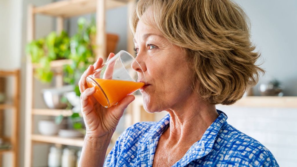 A woman in a blue shirt drinks a glass of orange juice in a kitchen with shelves and plants in the background.