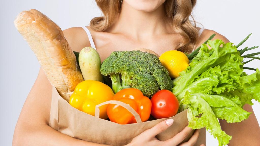 Woman holding a paper bag filled with fresh groceries including bread, peppers, lettuce, broccoli, and lemon.