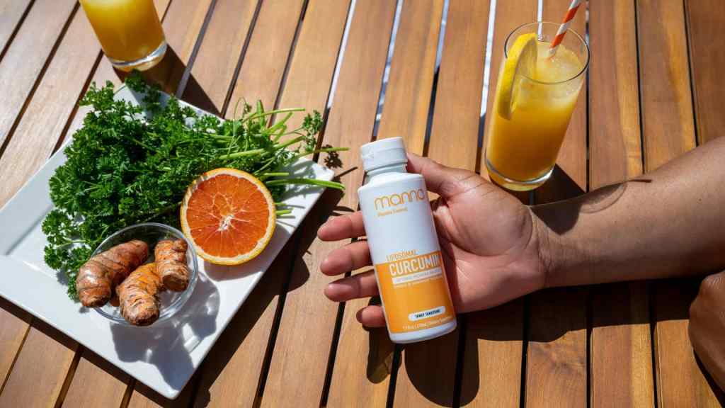 A hand holds a bottle labeled "Original Curcumin" on a wooden table with turmeric roots, parsley, orange slices on a plate, and a glass of orange juice.