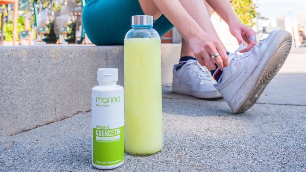 A person ties their shoe while sitting on the pavement beside a bottle labeled "Liposomal Quercetin" and a glass bottle filled with a yellow liquid.