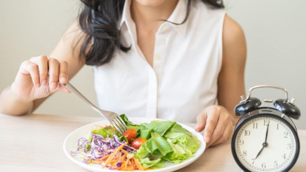 Woman eating a salad next to a clock showing 12 o'clock.