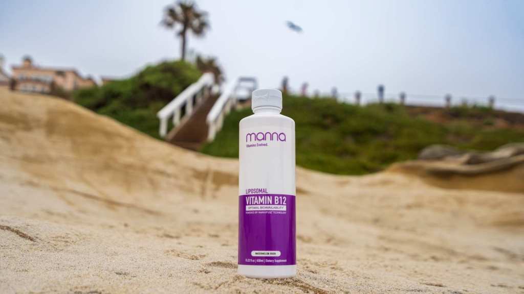 A bottle of vitamin B12 supplement stands on sandy ground with blurred greenery and steps in the background.