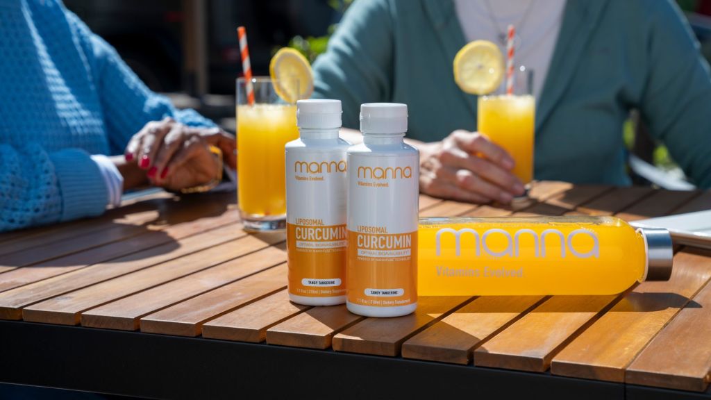 Two bottles of Manna Liposomal Curcumin stand on a wooden table with a Manna water bottle and two people drinking orange beverages in the background.