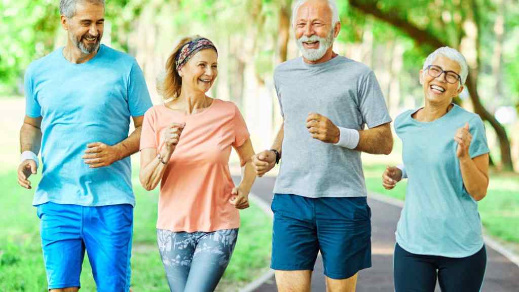 Four older adults, two men and two women, are jogging and smiling on a path in a park with trees and grass in the background.