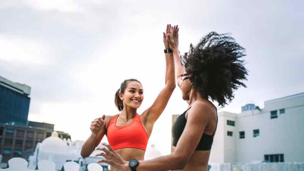 Two women in athletic wear high-fiving outdoors, with buildings in the background and a cloudy sky above.