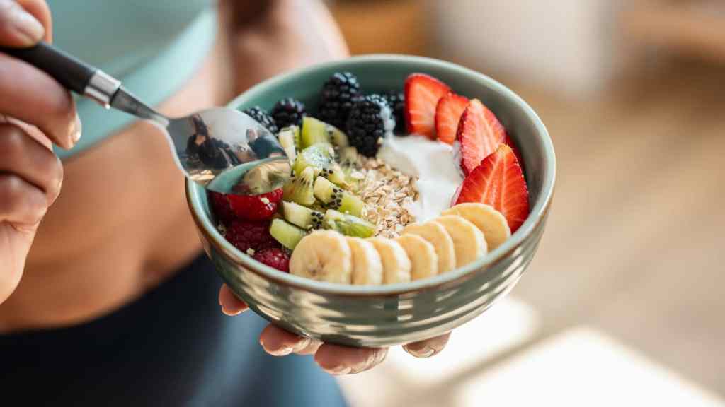 A person holds a bowl filled with sliced bananas, strawberries, kiwis, blackberries, granola, and yogurt, with a spoon ready to eat.