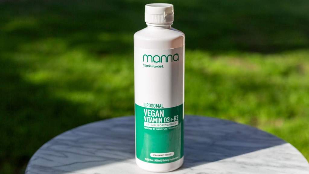 A bottle of Mana Liposomal Vegan Vitamin D3+K2 stands on a marble surface with grass in the background.