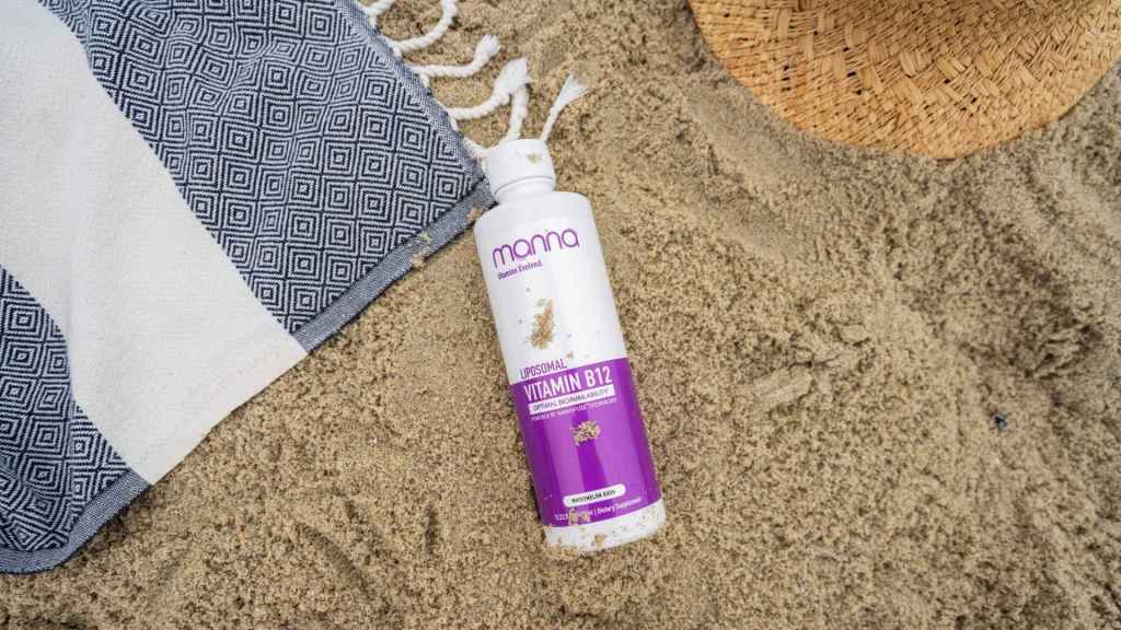 A bottle of purple Vitamin B12 lotion lies on sandy ground next to a woven mat and a straw hat.