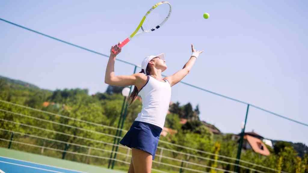 Person in athletic gear serves a tennis ball on an outdoor court, surrounded by greenery and a blue sky.