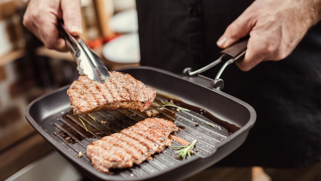 Person grilling two steaks in a grill pan with tongs, rosemary visible.