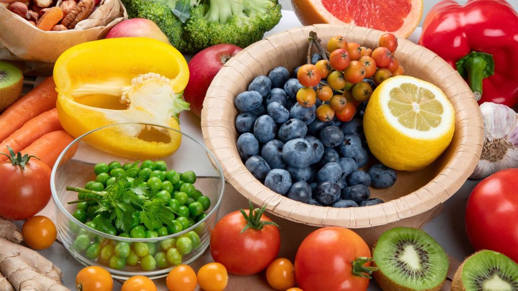 A variety of fruits and vegetables including broccoli, blueberries in a bowl, lemon, kiwi, tomatoes, carrots, bell pepper, peas, parsley, garlic, grapefruit, ginger, and nuts.