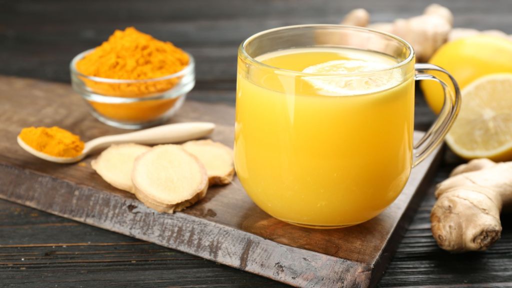 A glass of yellow turmeric and ginger beverage sits on a wooden board with a spoonful of turmeric powder, ginger slices, and a lemon nearby.