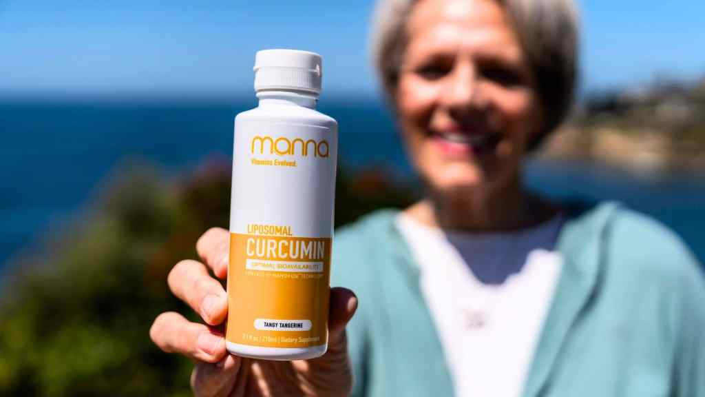 A person holding a bottle labeled "Liposomal Curcumin" by Mama, with the ocean and coastal landscape blurred in the background.