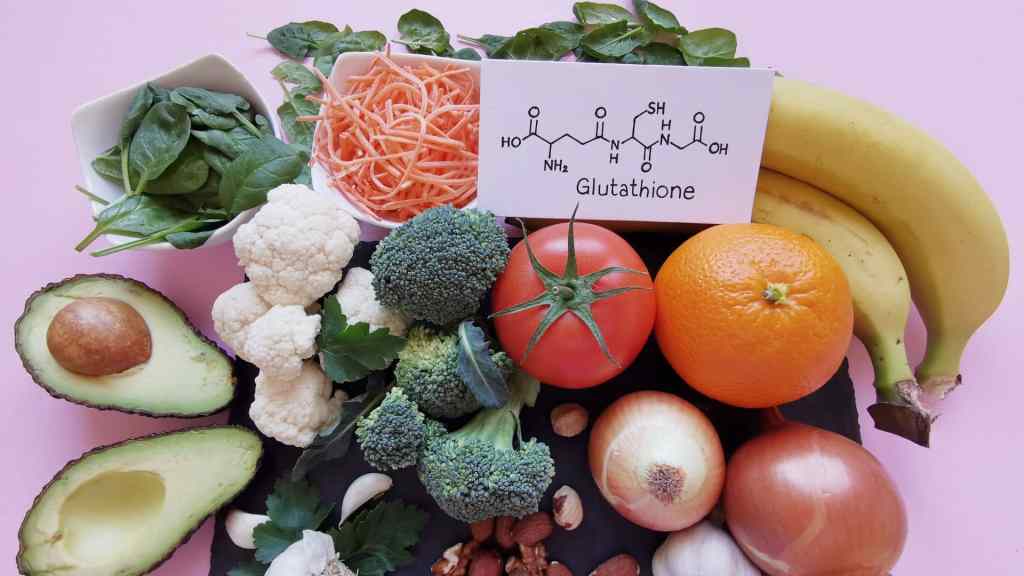 A variety of fruits and vegetables, including avocado, broccoli, tomato, and bananas, arranged around a card showing the chemical structure of glutathione, on a pink background.