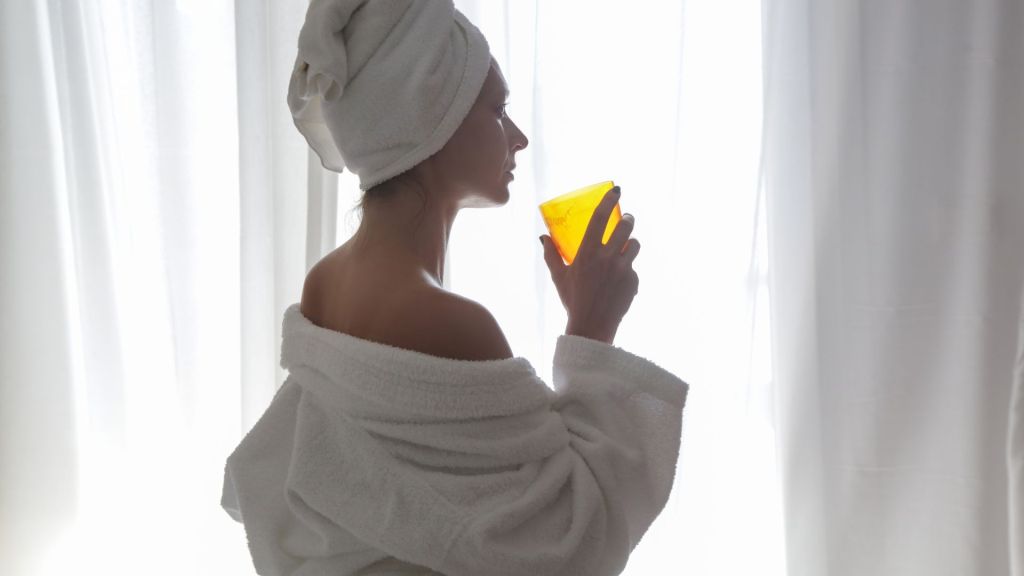 A person wearing a towel on their head and a white bathrobe is drinking from a yellow glass while standing in front of a window with sheer curtains.