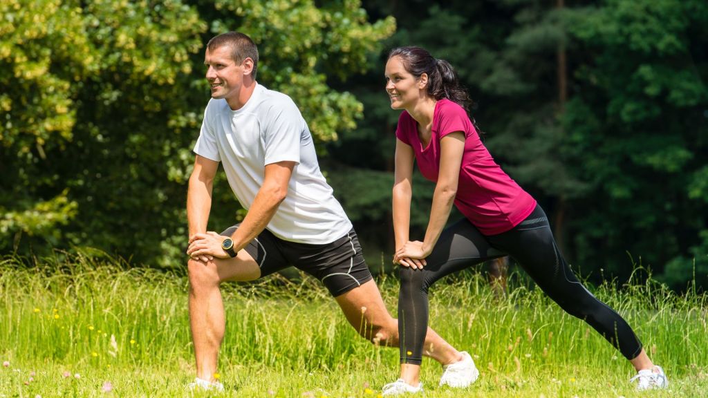 A man and woman in athletic attire perform lunges on a grassy field with trees in the background.