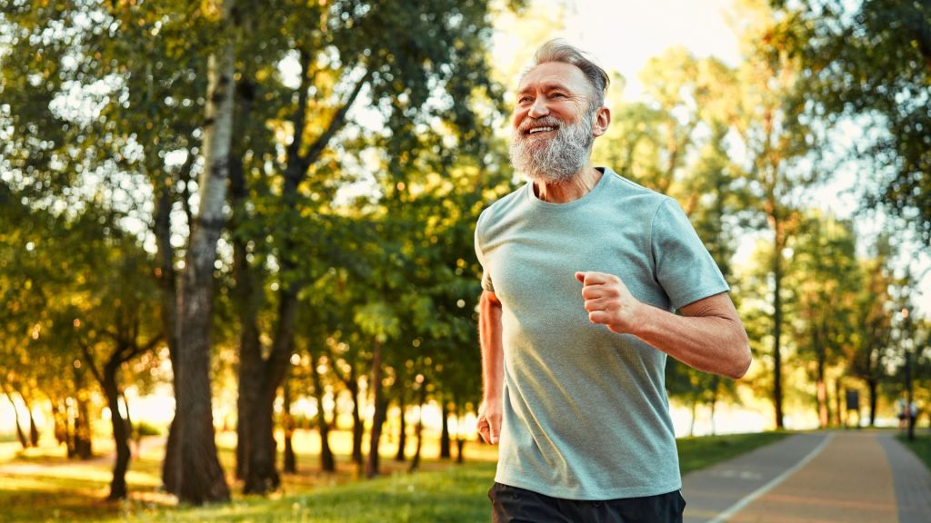 Elderly man with a gray beard jogging on a park path surrounded by green trees and sunlight.