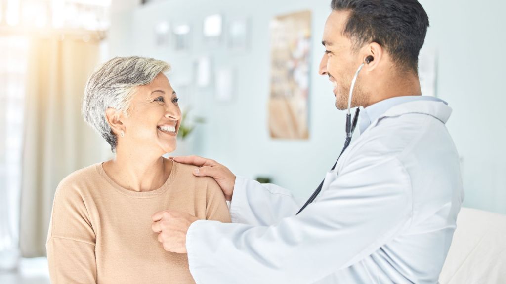 A doctor in a white coat, using a stethoscope, examines a smiling woman wearing a beige sweater in a bright room.