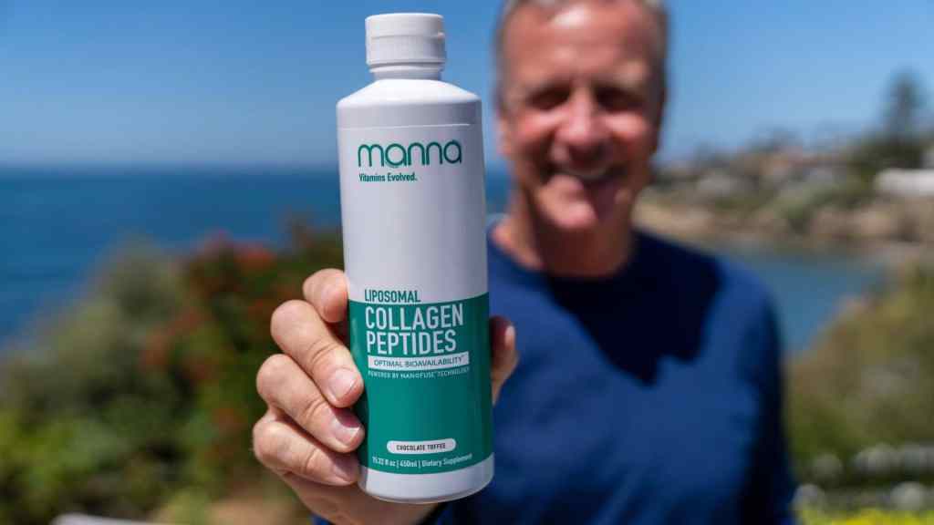 A man holds a bottle of liposomal collagen peptides supplement labeled "Manna Health" with an outdoor backdrop of the ocean and foliage.
