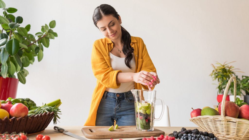 Woman at a counter surrounded by produce placing fruits into a blender.