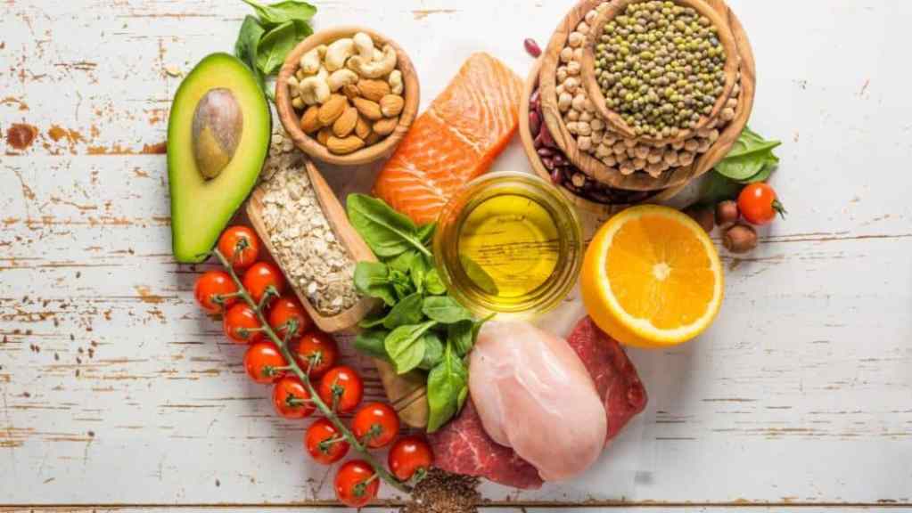 Assorted healthy foods including fruits, vegetables, nuts, grains, and lean meat displayed on a wooden surface.