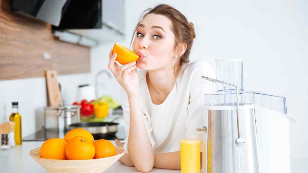 A woman leans on a kitchen counter, holding a half orange to her face. A bowl of oranges and a glass of juice are nearby, along with a juicer.