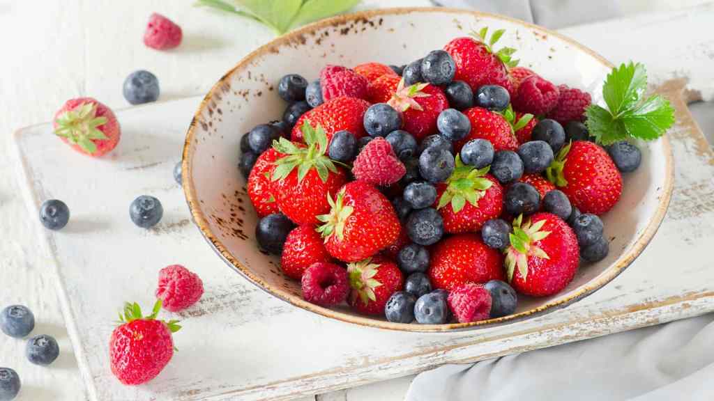 A bowl filled with fresh strawberries, blueberries, and raspberries on a rustic wooden board.