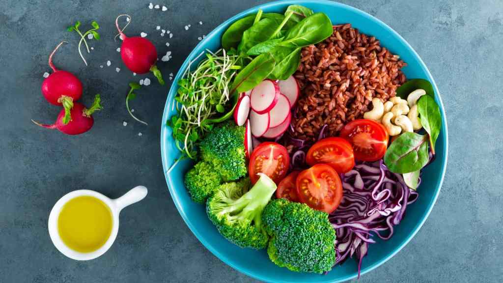 A blue bowl with red rice, spinach, broccoli, tomatoes, radishes, sprouts, purple cabbage, and cashews. A small jug of olive oil and radishes are beside the bowl.
