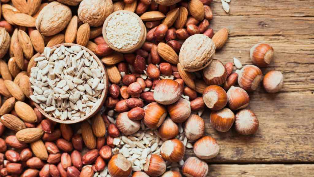 Assorted nuts and seeds, including almonds, walnuts, hazelnuts, peanuts, sunflower seeds, and sesame seeds, spread on a wooden surface.