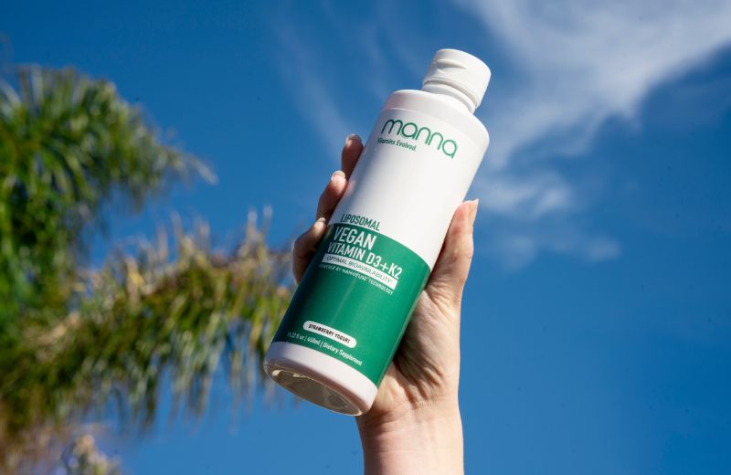 A hand holds a bottle labeled "Manna Vegan Vitamin D3 + K2" against a backdrop of blue sky and palm fronds.