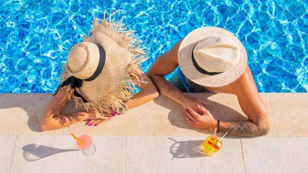 Two people relaxing by a swimming pool, with summer hats on and a tropical drink nearby.