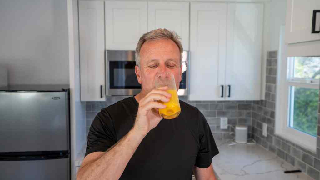 Man in a black shirt drinks orange juice in a modern kitchen with white cabinets and a microwave.