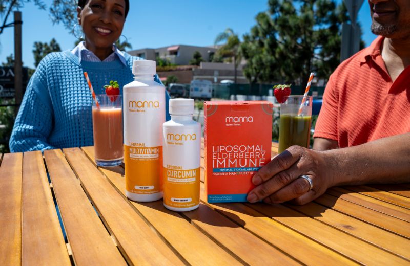 Two people sit at an outdoor table with three supplement bottles and two glasses of colorful smoothies. The people are out of focus in the background.