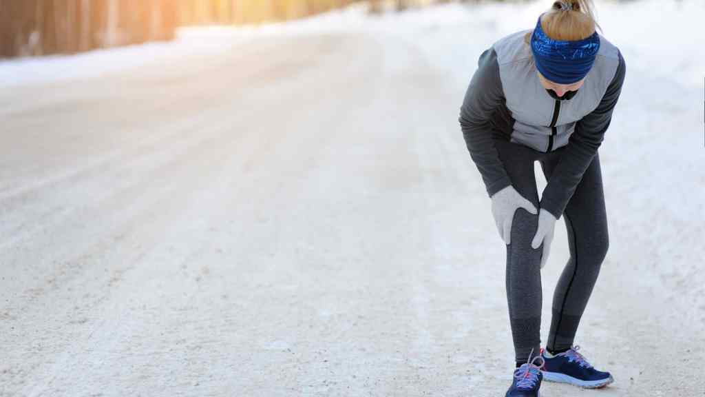 Person in winter gear holds their knee on a snowy road, appearing to be resting or in discomfort.