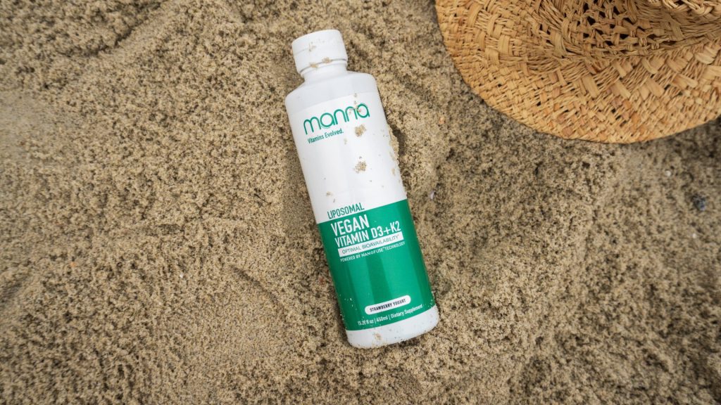 A bottle of Manna Liposomal Vegan Vitamin D3+K2 is lying on the sand next to a straw hat.