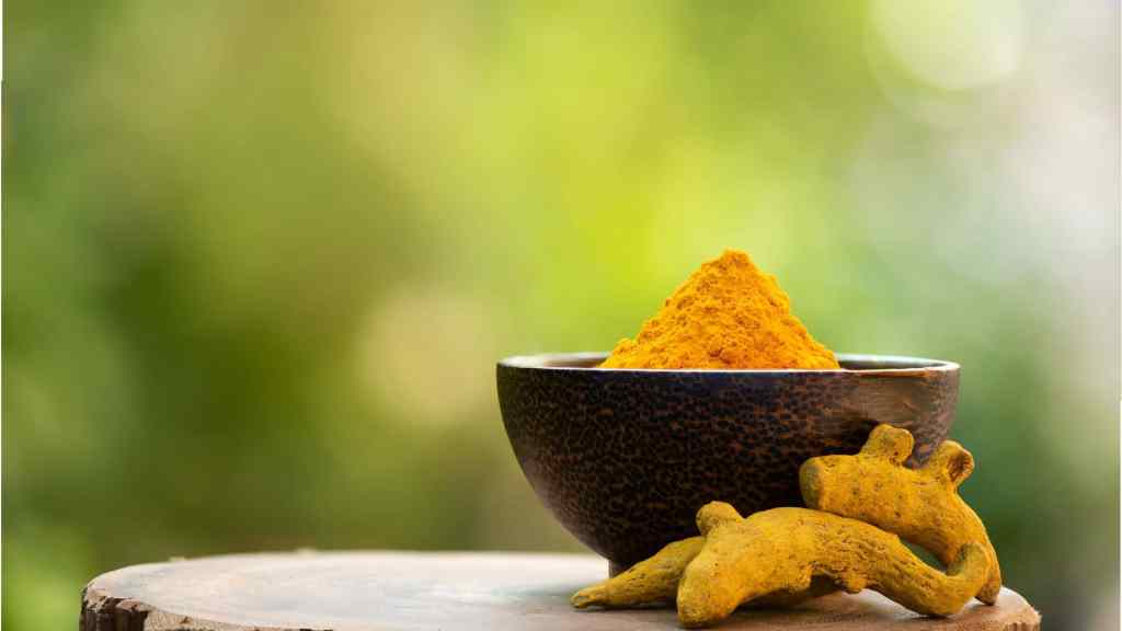 A ceramic bowl filled with ground turmeric sits on a wooden surface, with turmeric roots beside it, against a blurred green background.