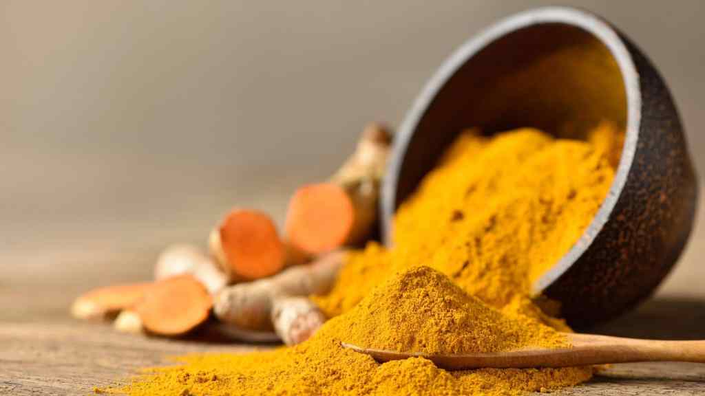 A wooden bowl spills turmeric powder onto a wooden surface, with a few turmeric roots in the background.
