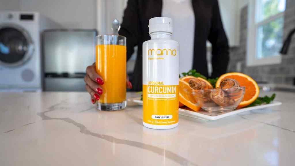 A bottle of liposomal curcumin supplement on a kitchen counter with a person holding a glass of orange juice, a plate of orange slices, and a small bowl in the background.