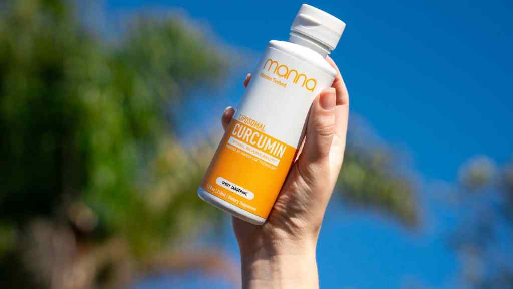 A hand holds a bottle of Manna Liposomal Curcumin supplement against a blue sky and blurred greenery background.
