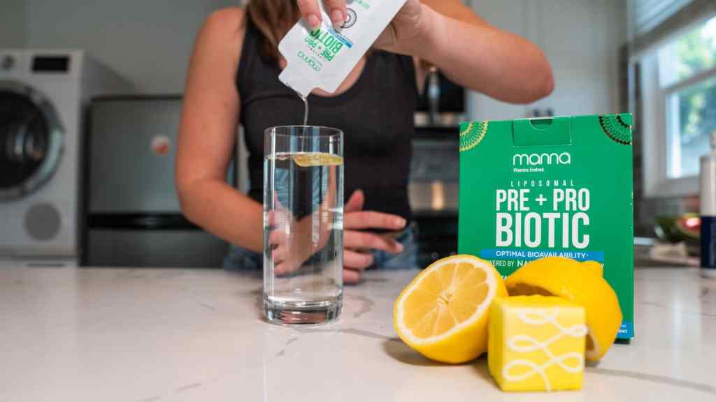 A person pours a liquid supplement from a packet into a glass of water next to a box labeled "Pre + Pro Biotic" and sliced lemons on a kitchen counter.