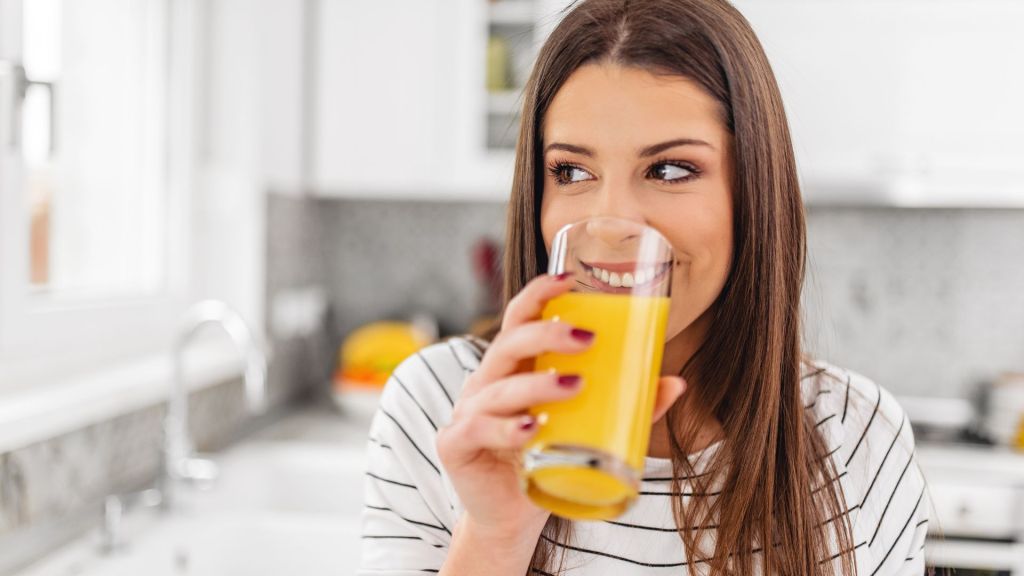 A woman in a striped shirt is smiling while drinking a glass of orange juice in a kitchen setting.