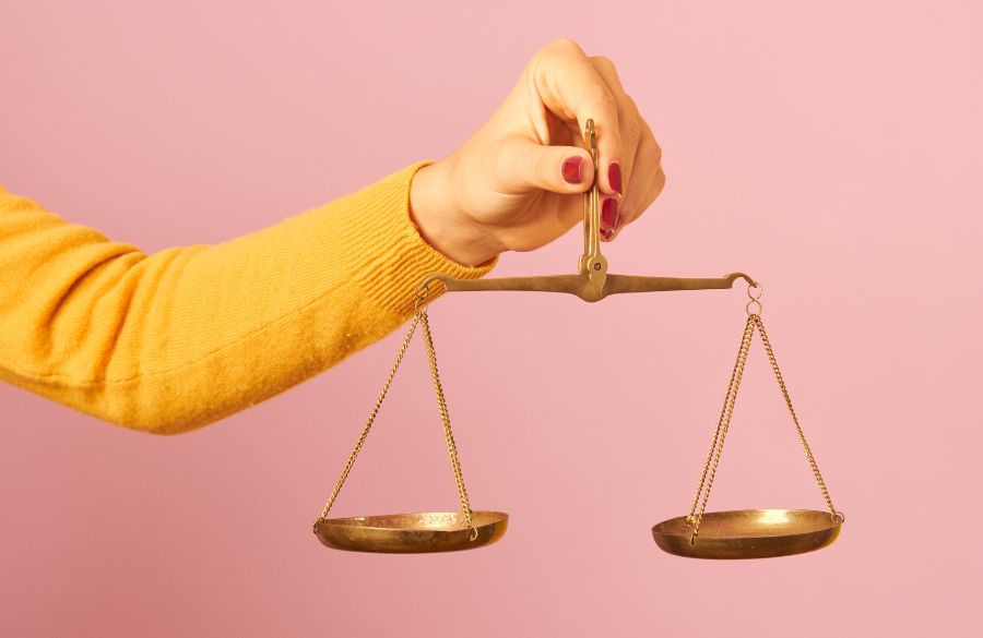 A person in a yellow sweater holding a small brass balance scale on a pink background.