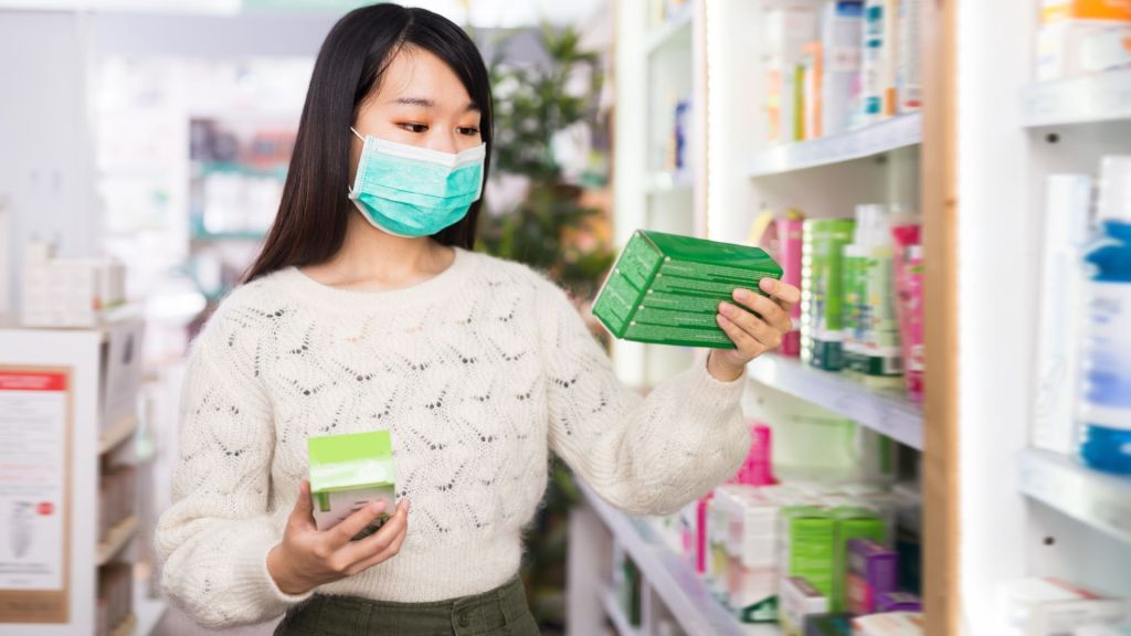 Woman wearing a mask holds two green boxes while examining products on a pharmacy shelf.