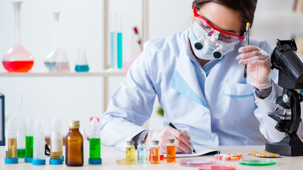 A scientist in a lab coat writes notes. Various lab equipment and colorful liquid-filled containers are on the table alongside a microscope.